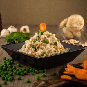 Italiana rižoto sa graškom, šargarepom i piletinom poslužen u crnom tanjiru, sa svežim sastojcima u pozadini